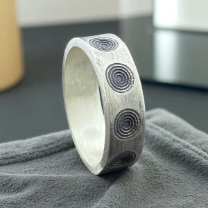 Könnte beinhalten: Ein silberner Ring mit einer Reihe von eingravierten konzentrischen Kreisen. Der Ring hat ein breites Band und ein gebürstetes Metall-Finish. Das Design wiederholt sich um den Umfang und erzeugt ein einzigartiges Muster.