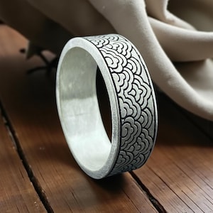 Może przedstawiać: Srebrny pierścionek z czarnym, powtarzającym się wzorem fal. Pierścionek jest gruby i ma gładkie, wypolerowane wnętrze. Pierścionek jest prezentowany na drewnianej powierzchni z tkaniną w neutralnym kolorze w tle.
