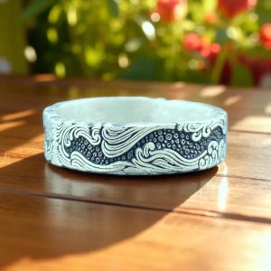 May include: A silver-colored ring with an intricate wave pattern design. The ring has a wide band and a textured surface. The wave pattern is detailed with small, circular accents. The ring is displayed on a wooden surface with natural light.