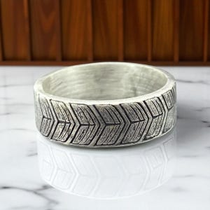 May include: A silver ring with a textured chevron pattern around the band. The ring has a brushed metal finish and a wide band design. The ring is displayed on a white marble surface.