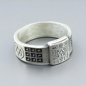 May include: Silver-toned ring with engraved geometric and symbolic designs. The ring features a square panel with a grid of symbols, and other panels with intricate patterns including stars, eyes, and suns. The ring has a wide band and a matte finish.