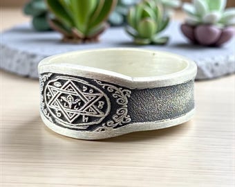 May include: A cream-colored ring with a detailed, dark gray band featuring a six-pointed star and other symbols. The ring sits on a wooden surface, with succulents in the background.