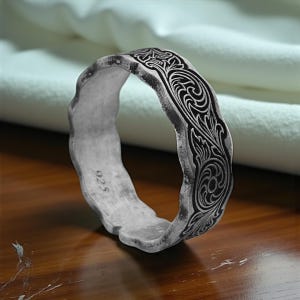 May include: A silver ring with an intricate floral design. The ring has a textured, wavy edge and the number "925" is stamped on the inside. The engraved pattern is dark against the silver.
