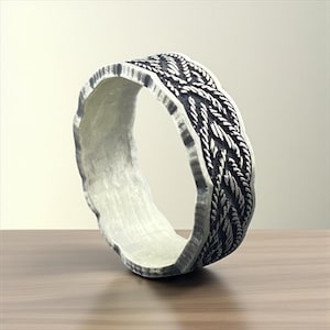 Puede incluir: Un anillo plateado con un diseño trenzado texturizado alrededor de la banda. El anillo tiene un borde áspero e irregular y un interior liso y de color claro. El anillo se muestra sobre una superficie de madera.
