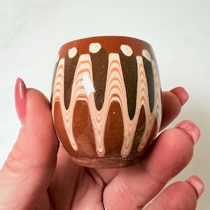 May include: A small, rounded ceramic cup featuring a marbled pattern of deep brown, terracotta, and cream-colored swirls. The design consists of vertical, wave-like stripes that extend from the rim toward the base, accented by small cream dots.