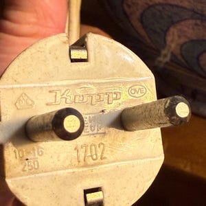 May include: Close-up of a cream-colored electrical plug with two round prongs and a rectangular ground pin. The plug has the text "Kopp" and the number "1702" printed on it. The plug is connected to a white electrical cord.