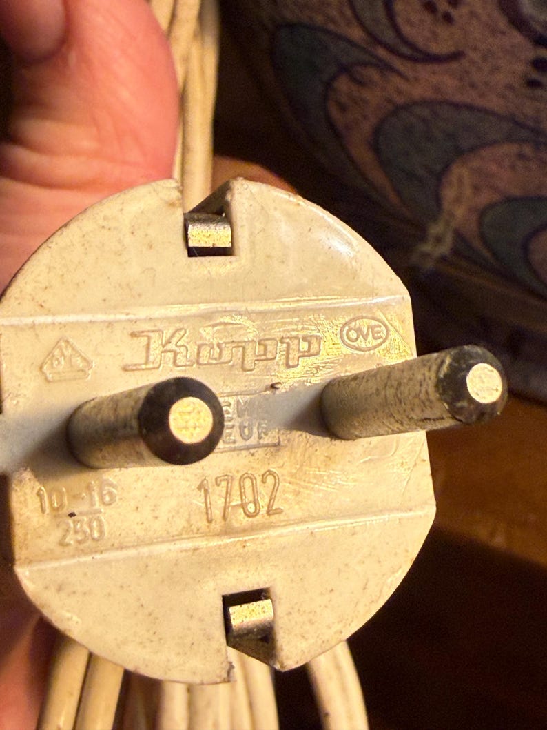 May include: Close-up of a cream-colored electrical plug with two round prongs and a rectangular ground pin. The plug has the text "Kopp" and the number "1702" printed on it. The plug is connected to a white electrical cord.