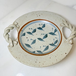 May include: A speckled ceramic serving platter with two twisted rope handles. The center features a blue and brown design with blue leaf motifs. The rim has a brown and blue border. The overall color palette is neutral with brown and blue accents.