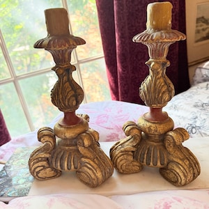 May include: A pair of matching candle holders featuring a gold-toned finish with distressed red accents. Each holder has a tiered base with scroll-like feet, a central bulbous stem with carved leaf details, and a fluted top cup for a candle.