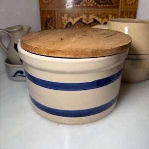 May include: A cylindrical cream-colored ceramic crock featuring two horizontal navy blue stripes encircling the body. The container is topped with a circular, flat, light brown wooden lid that has a visible grain and a small center hole.
