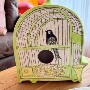 May include: A ceramic birdcage in shades of green and white, featuring a black and white bird illustration. The cage has a circular opening and decorative floral accents. The cage is on a wooden surface.