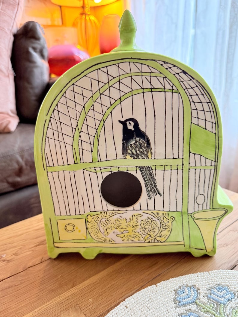 May include: A ceramic birdcage in shades of green and white, featuring a black and white bird illustration. The cage has a circular opening and decorative floral accents. The cage is on a wooden surface.