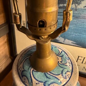 May include: Close-up of a ceramic lamp base with a brass lamp holder. The base is white with a blue and teal floral pattern. The lamp's metal components have a gold finish. The lamp is on a wooden surface.