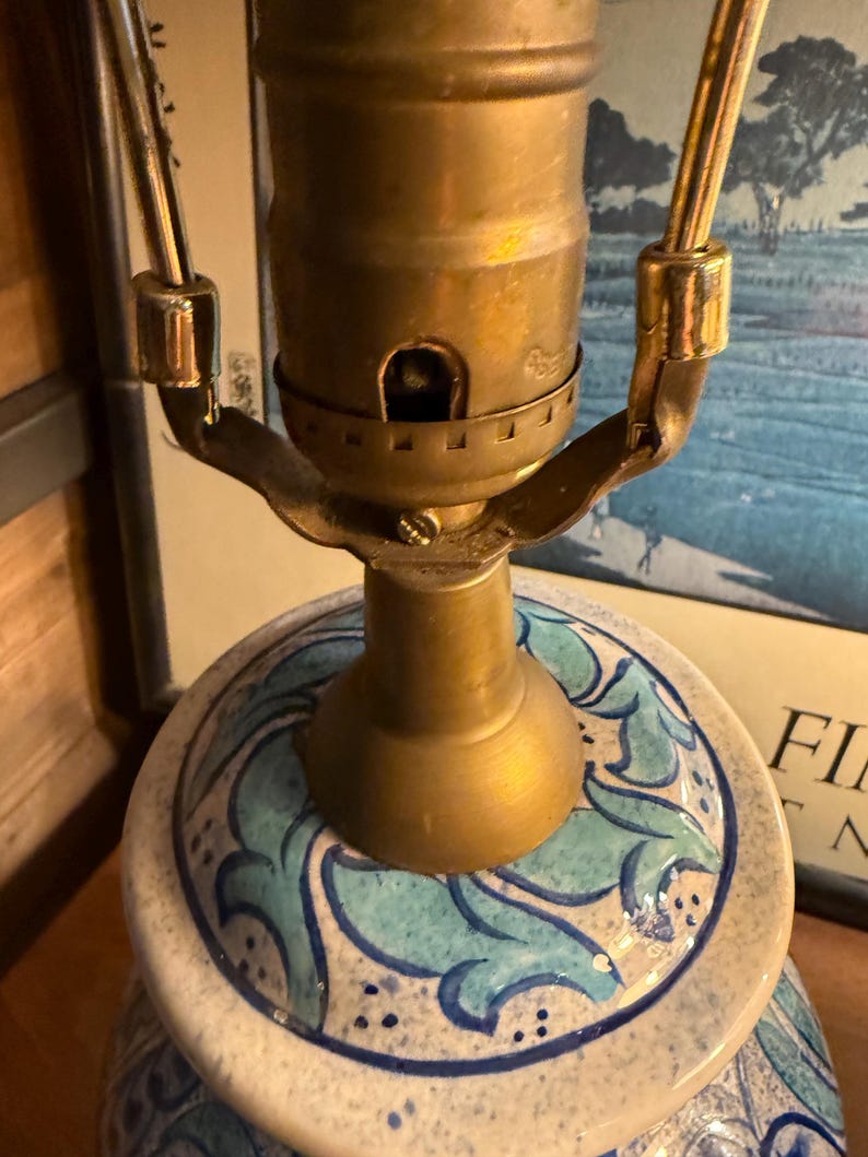 May include: Close-up of a ceramic lamp base with a brass lamp holder. The base is white with a blue and teal floral pattern. The lamp's metal components have a gold finish. The lamp is on a wooden surface.