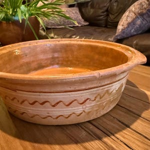 May include: A large, round, earthenware bowl with a wide rim and two small handles. The bowl is a warm terracotta color with a decorative wavy pattern around the lower portion. The bowl sits on a wooden surface.