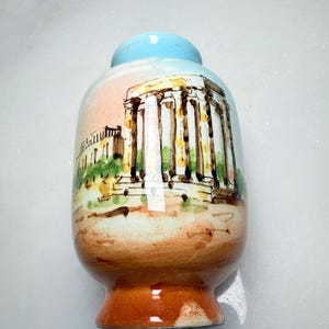 May include: A small ceramic vase with a light blue neck and a rounded body featuring a hand-painted illustration of a classical temple with columns. The base is a warm orange-brown, and the surface displays a fine crackle glaze finish throughout.