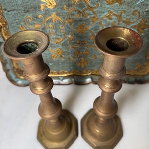 May include: Two antique brass candlesticks. Each candlestick has a flared top, a bulbous midsection, and a hexagonal base. The brass has a warm, aged patina. The background features a teal and gold decorative tray.