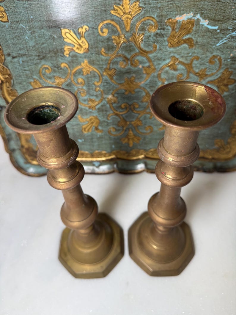 May include: Two antique brass candlesticks. Each candlestick has a flared top, a bulbous midsection, and a hexagonal base. The brass has a warm, aged patina. The background features a teal and gold decorative tray.