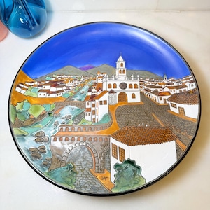 May include: A round decorative ceramic plate featuring a hand-painted scene of a town with white buildings, terracotta roofs, a stone bridge over a river, and a central church with a clock tower, all set against a vibrant blue sky and mountains.