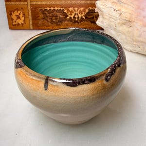 May include: A ceramic bowl with a vibrant turquoise interior and a smooth, light tan exterior featuring a crackled glaze finish. The rim is accented with dark brown, drip-like glaze patterns that extend slightly down the outer surface of the bowl.