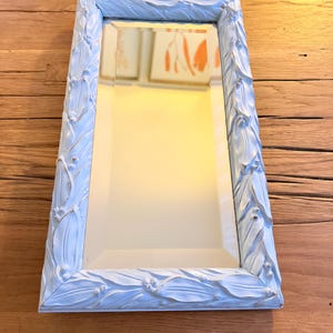 May include: Rectangular mirror with a light blue, ornate frame featuring a leaf and berry design. The mirror reflects a warm, yellow-toned light. The frame's details add a vintage or antique aesthetic to the piece.