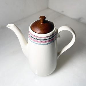 May include: White ceramic teapot with a wooden lid and handle. The teapot features a decorative band of pink, white, and blue geometric patterns around the middle. The spout is long and curved.