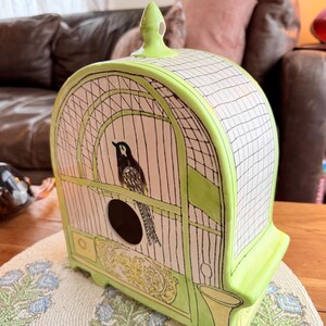 May include: A ceramic birdcage with a black and white bird inside. The cage is painted in light green and white, with a black grid pattern. The base has a decorative yellow design. The birdcage is sitting on a beaded placemat.