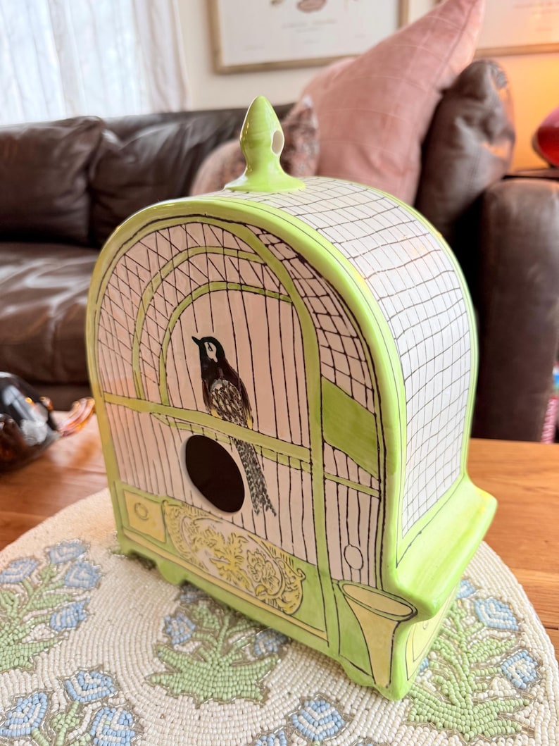 May include: A ceramic birdcage with a black and white bird inside. The cage is painted in light green and white, with a black grid pattern. The base has a decorative yellow design. The birdcage is sitting on a beaded placemat.