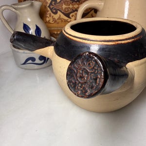 May include: A round, beige ceramic teapot with a dark brown glazed band around the top and a matching dark brown handle featuring a circular embossed seal. The teapot has a short, dark brown spout and a wide opening at the top.