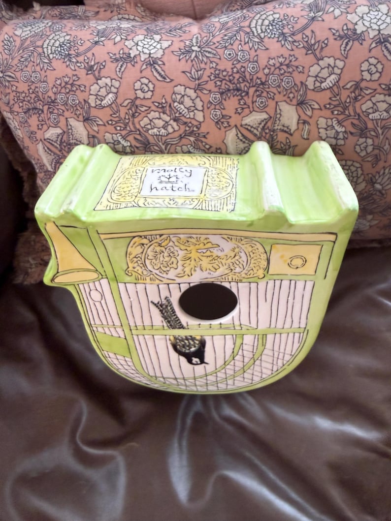 May include: A decorative birdhouse in a unique carriage shape, painted in light green and cream. The birdhouse features a black bird illustration, a round entry hole, and the text "molly hatch".