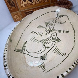 May include: Oval ceramic serving dish with a cream-colored base and a hand-painted design of two fish in green. The rim is decorated with a series of dark green dashes. A woven basket and a wooden decorative box are in the background.
