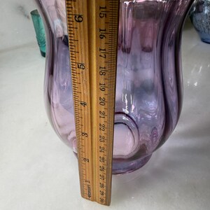 Peut inclure: Un vase en verre de couleur lavande avec un bord festonn&eacute;. Le vase a une forme bulbeuse et mesure environ 20 cm de hauteur. Une r&egrave;gle en bois est plac&eacute;e &agrave; c&ocirc;t&eacute; du vase pour la mesure.