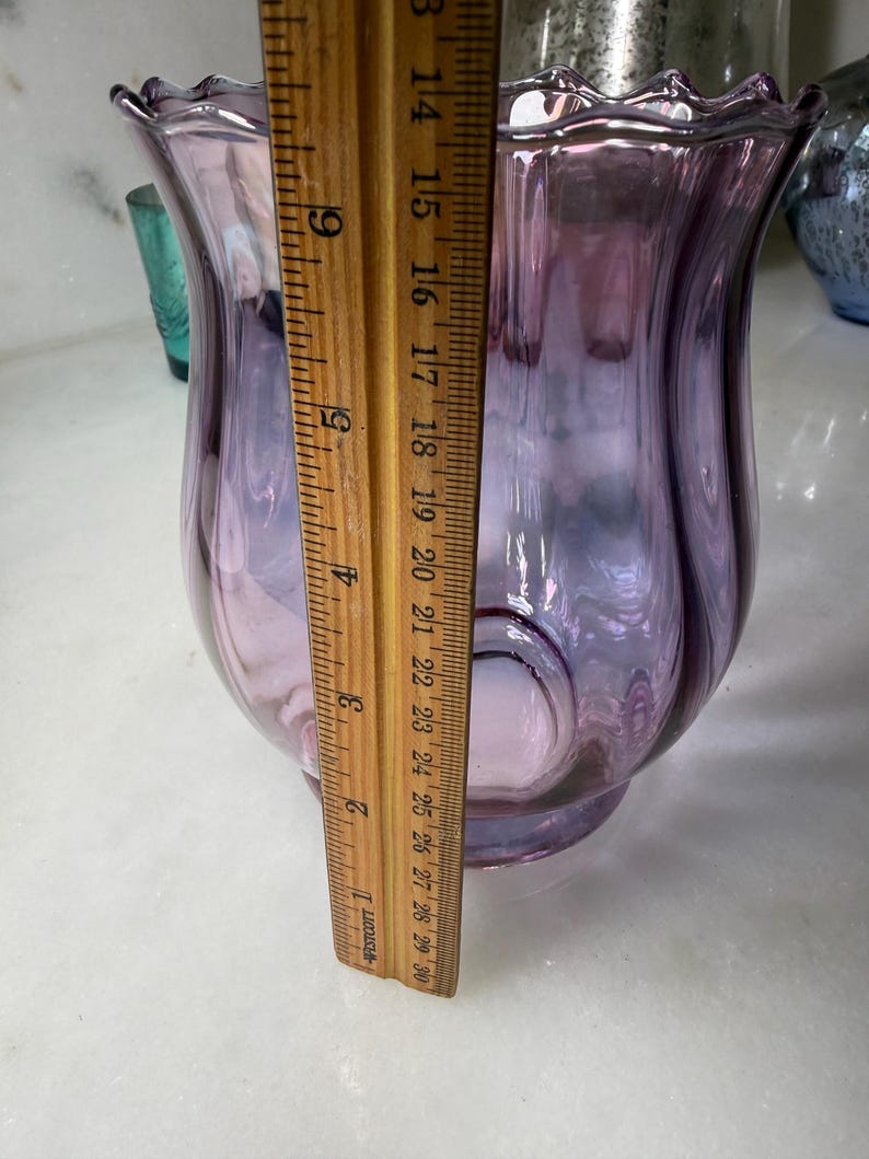 Peut inclure: Un vase en verre de couleur lavande avec un bord festonn&eacute;. Le vase a une forme bulbeuse et mesure environ 20 cm de hauteur. Une r&egrave;gle en bois est plac&eacute;e &agrave; c&ocirc;t&eacute; du vase pour la mesure.