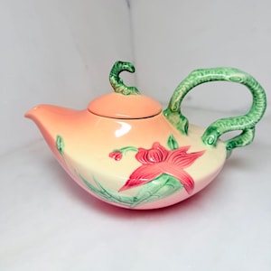 May include: A ceramic teapot with a peach-colored lid and body, featuring a large pink flower design with green leaves. The handle and spout are green, and the teapot has a curved shape. A decorative floral teapot.