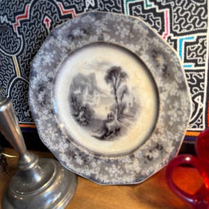 May include: Antique octagonal plate with a black and white landscape scene in the center. The plate has a gray floral border. A silver candlestick and a red glass pitcher are in the foreground.