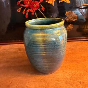 May include: A ceramic vase with a rounded body and a wide opening. The exterior features a mottled glaze in shades of deep blue, teal, and green, with a distinct yellowish-green rim. The surface shows subtle horizontal ridges from the pottery process.