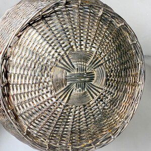 Puede incluir: Primer plano de una cesta tejida de color plateado. La cesta tiene forma redonda con un patr&oacute;n tejido apretado. El tejido crea una superficie texturizada, y la cesta parece ser de metal.