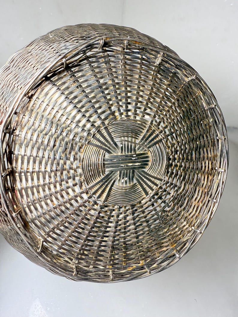 Puede incluir: Primer plano de una cesta tejida de color plateado. La cesta tiene forma redonda con un patr&oacute;n tejido apretado. El tejido crea una superficie texturizada, y la cesta parece ser de metal.