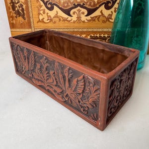 May include: A rectangular, brown ceramic planter with detailed relief carvings of birds and foliage. The planter has a reddish-brown exterior and a darker brown interior. A green glass bottle is visible in the background.