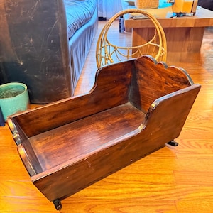 May include: A vintage wooden cradle with a dark brown finish. The cradle has a rectangular shape with curved sides and decorative detailing on the head and foot. It sits on small, rounded feet.