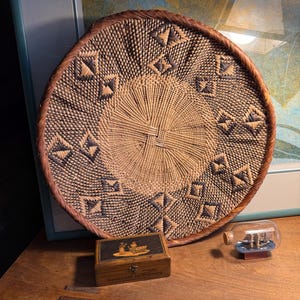 May include: A woven basket with a circular design, featuring geometric patterns in shades of brown and dark blue. A small wooden box with a decorative top and the text "Souvenir de Canaro" is next to the basket. A ship in a bottle is also present.