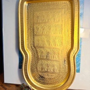May include: A gold-coloured rectangular tray with an embossed design featuring figures and patterns. The tray has a curved handle area and a decorative border. A small, detailed figurine of a horse and a ship in a bottle are also visible.