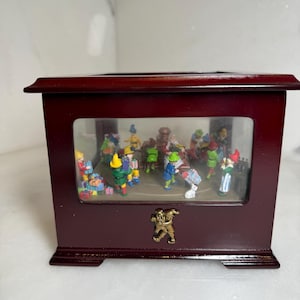 May include: A dark red wooden music box with a clear front panel, showcasing miniature figures in colorful outfits. A gold-colored emblem is on the front. The box is rectangular with a decorative base.