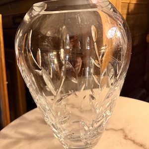 May include: A clear, cut-glass vase with a wide, rounded body and a slightly flared rim. The vase features an etched leaf and stem design. The vase is sitting on a marble surface.