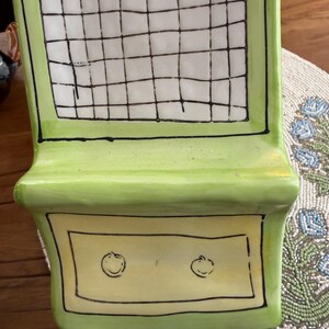 May include: A decorative, ceramic shelf in shades of green and yellow. The shelf features a grid pattern on the top section and two small, stylized fruit designs on the lower section. Black outlines define the edges and details.