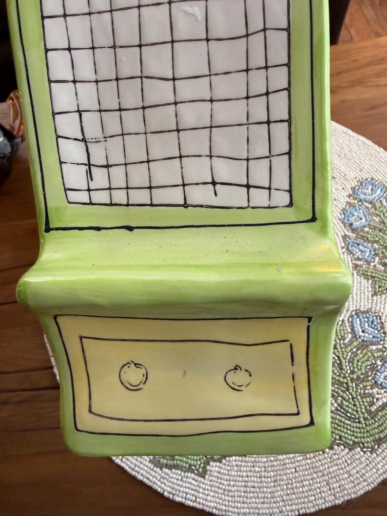 May include: A decorative, ceramic shelf in shades of green and yellow. The shelf features a grid pattern on the top section and two small, stylized fruit designs on the lower section. Black outlines define the edges and details.