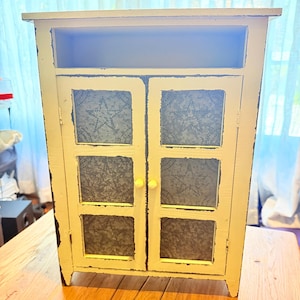May include: A small, rectangular white cabinet with a distressed finish, featuring two doors with three inset panels each. The panels are made of gray metal with a punched star pattern. The cabinet includes a single open shelf at the top.