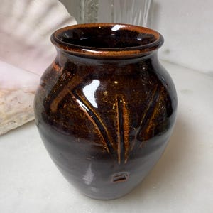 May include: A small, rounded ceramic vase with a glossy, dark brown glaze. The surface features subtle, carved vertical lines radiating from the center and a small, square-shaped indentation near the base. The rim is smooth and slightly flared.