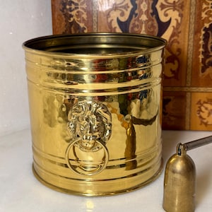 May include: A gold-colored metal bucket with a hammered texture. The bucket has a lion's head with a ring handle. A small, bell-shaped object is next to the bucket.