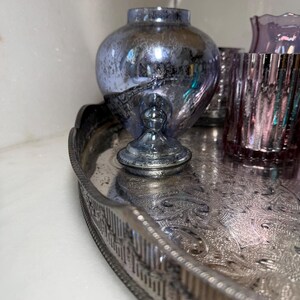 Peut inclure: Un plateau argent&eacute; orn&eacute; contient une collection d'objets en verre d&eacute;coratifs. Un grand vase bleu&acirc;tre de forme bulbeuse repose sur un pi&eacute;destal. Plusieurs verres roses et violets sont &eacute;galement sur le plateau.